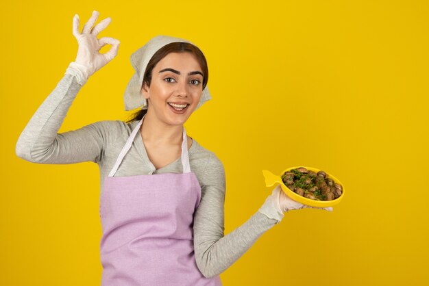 Young female cook in apron holding plate of fried mushrooms and showing ok sign.