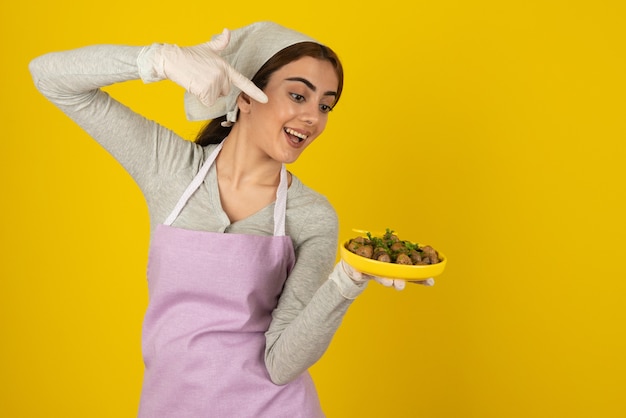 노란색 벽 위에 튀긴 버섯 접시를 들고 앞치마에 젊은 여성 요리사.