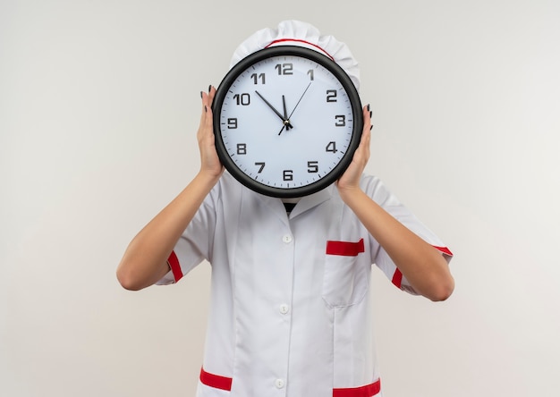 Young female cook in chef uniform holding and hiding behind clock isolated on white background