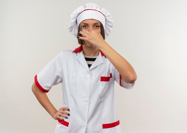 Giovane cuoco femminile in uniforme del cuoco unico che tiene il naso con la mano sulla vita che guarda l'obbiettivo isolato su fondo bianco con lo spazio della copia