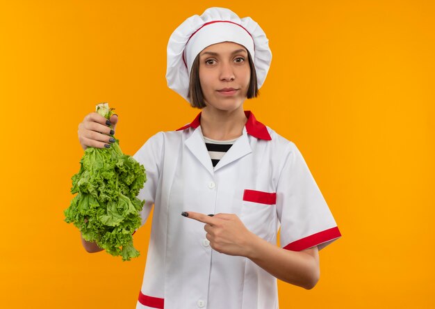 Young female cook in chef uniform holding and pointing at lettuce and looking at camera isolated on orange background