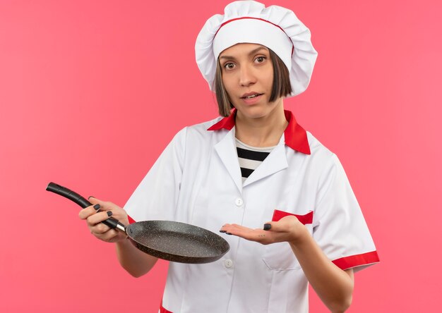 Young female cook in chef uniform holding and pointing with hand at frying pan looking at camera isolated on pink background