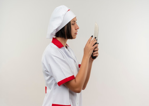 Молодая женщина-повар в униформе шеф-повара, стоя в профиль, держа и глядя на нож, изолированные на белом фоне с копией пространства