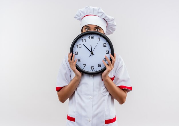 young female cook wearing chef uniform covered face with wall clock on isolated white wall with copy space