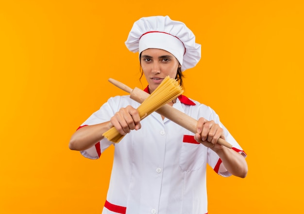 요리사 유니폼을 입고 복사 공간이 격리 된 노란색 벽에 롤링 핀으로 스파게티를 건너는 젊은 여성 요리사