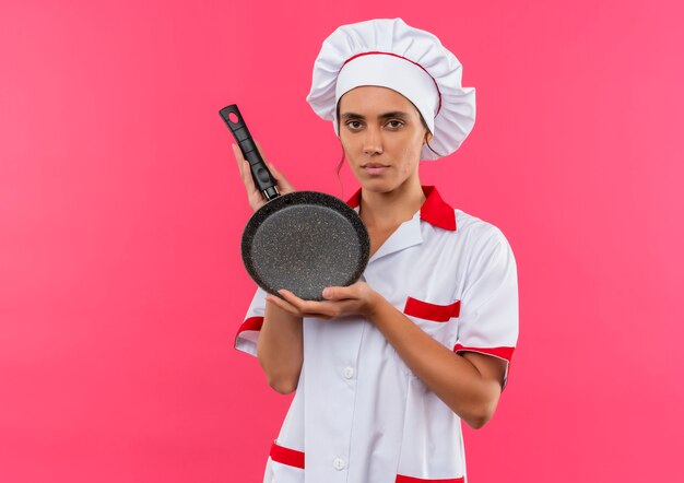  young female cook wearing chef uniform holding frying plate on isolated pink wall with copy space