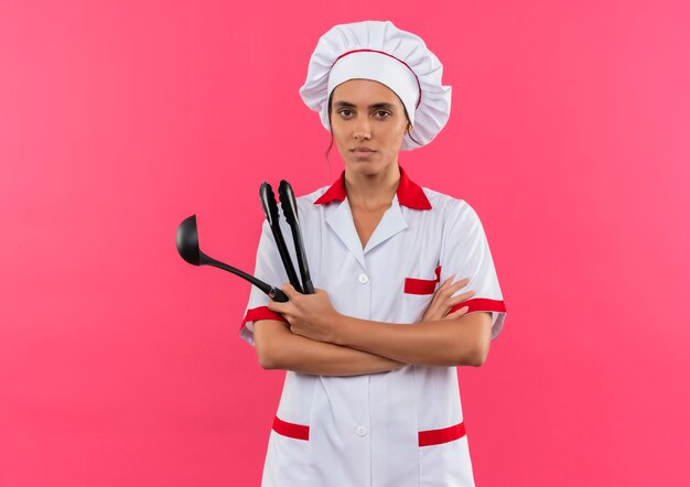  young female cook wearing chef uniform holding ladle and crossing hands on isolated pink wall with copy space