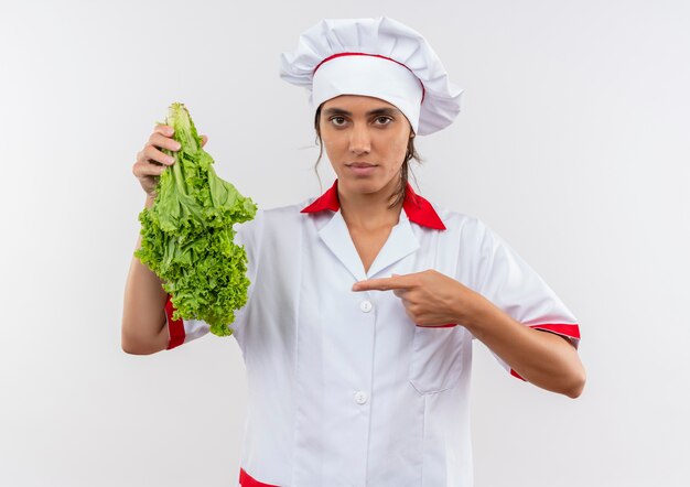 young female cook wearing chef uniform holding and points to salad on isolated white wall with copy space