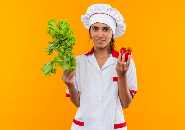 молодая женщина-повар в униформе шеф-повара держит салат и перец на изолированной желтой стене с копией пространства