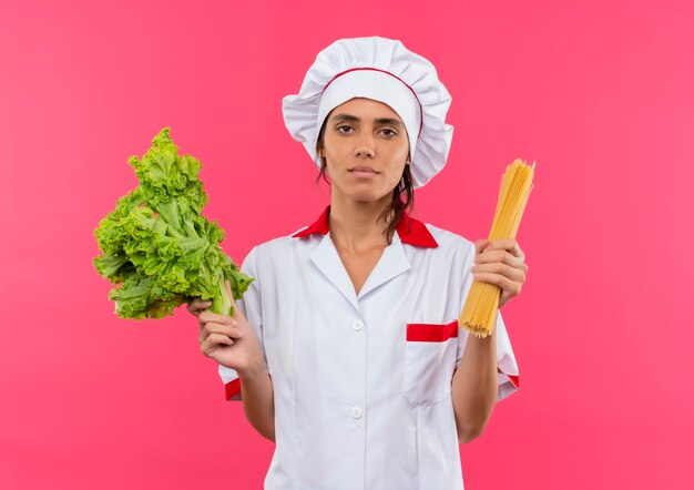 молодая женщина-повар в униформе шеф-повара держит спагетти и салат на изолированной розовой стене с копией пространства