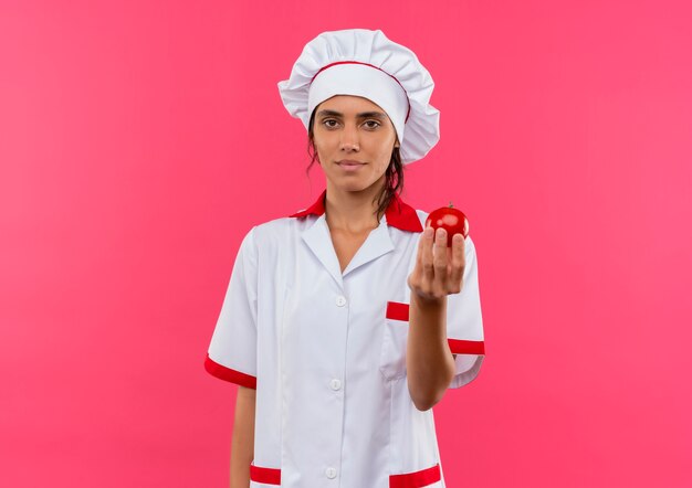  young female cook wearing chef uniform holding tomato on isolated pink wall with copy space