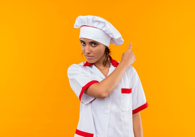 Free photo young female cook wearing chef uniform points to back on isolated yellow wall with copy space