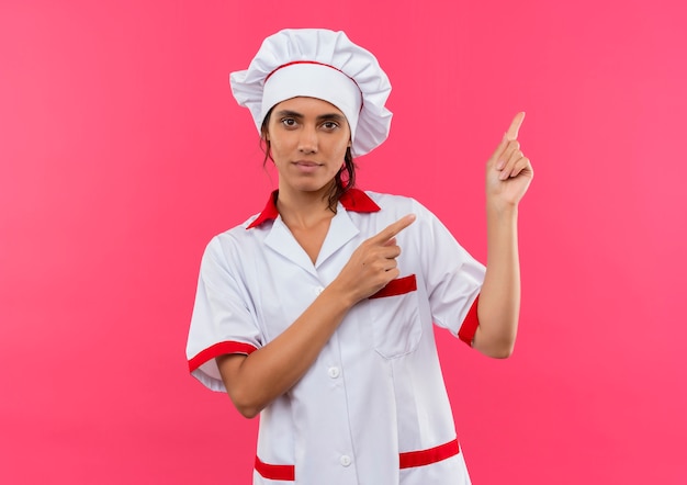  young female cook wearing chef uniform points to side on isolated pink wall with copy space