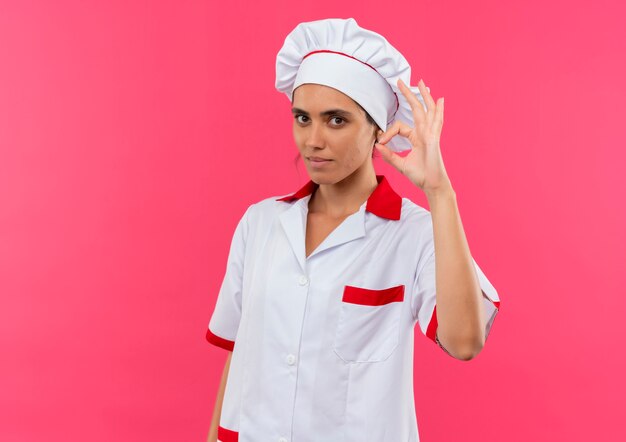 コピースペースと孤立したピンクの壁に大丈夫なジェスチャーを示すシェフの制服を着た若い女性料理人
