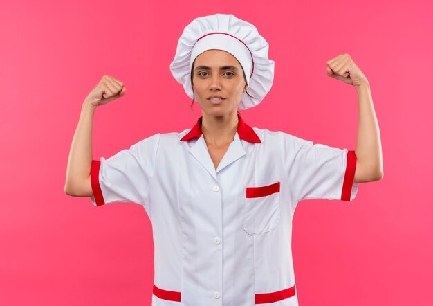 복사 공간이 격리 된 분홍색 벽에 강한 제스처를 보여주는 요리사 유니폼을 입고 젊은 여성 요리사