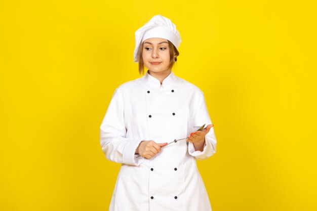 молодая женщина-повар в белом кулинарном костюме и белой кепке позирует недовольно держа серебряную ложку