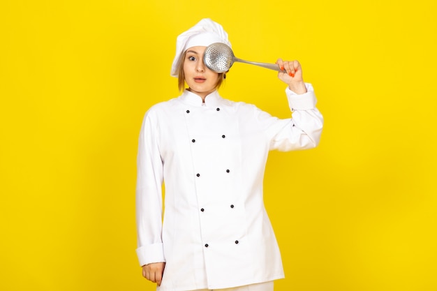 若い女性が白いクックスーツと彼女の目を覆っている銀のスプーンを保持している思考をポーズホワイトキャップで調理