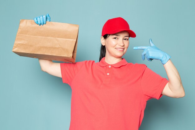 青い壁にボックスを保持しているピンクのtシャツの赤い帽子青い手袋の若い女性の宅配便