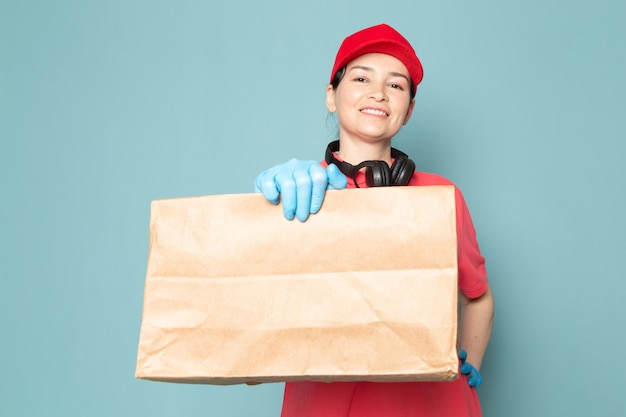 young female courier in pink t-shirt red cap blue gloves holding box on the blue wall
