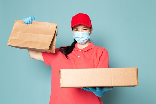 분홍색 티셔츠에 젊은 여성 택배 빨간 모자 파란색 무균 마스크 파란색 장갑 파란색 벽에 상자를 들고