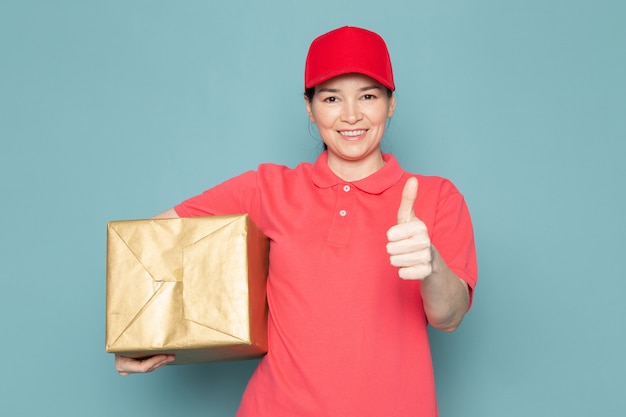 young female courier in pink t-shirt red cap holding box on the blue wall