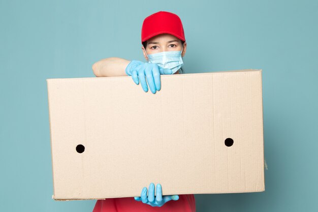 Free photo young female courier in pink t-shirt red cap holding box on the blue wall