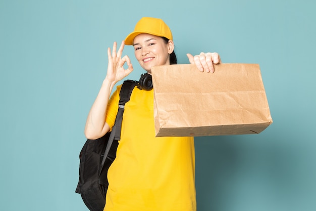 青い壁にボックスを保持している黄色のtシャツイエローキャップの若い女性宅配便