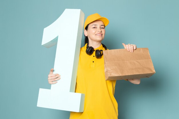 молодая женщина-курьер в желтой футболке желтая кепка держит коробку на синей стене