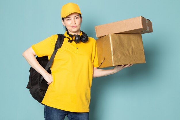 young female courier in yellow t-shirt yellow cap holding box on the blue wall