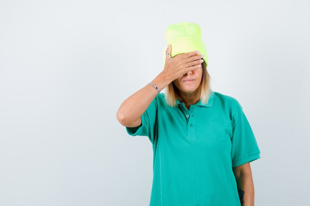 Free photo young female covering eyes with hand in polo t-shirt, beanie and looking curious , front view.