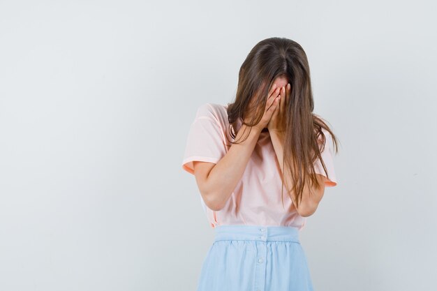 Young female covering face with hands in t-shirt, skirt and looking sorrowful. front view.