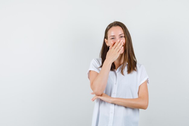 Young female covering hand on mouth while secretly laughing in white blouse and looking merry.