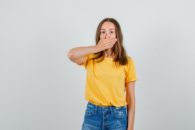 Young female covering mouth with hand in t-shirt, shorts and looking silent. front view.