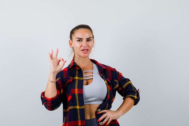 Giovane donna in crop top, camicia a scacchi, pantaloni che mostrano un gesto ok mentre si tiene la mano sull'anca e sembrano fiduciosi, vista frontale.