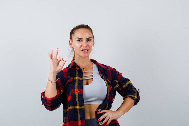 Young female in crop top, checkered shirt, pants showing ok gesture while keeping hand on hip and looking confident , front view.