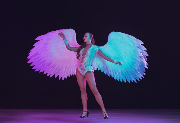 Молодая танцовщица с крыльями белого ангела в фиолетовом синем неоновом свете на черной стене.