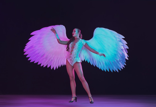 검은 벽에 보라색 파란색 네온 불빛에 흰색 천사의 날개를 가진 젊은 여성 댄서.