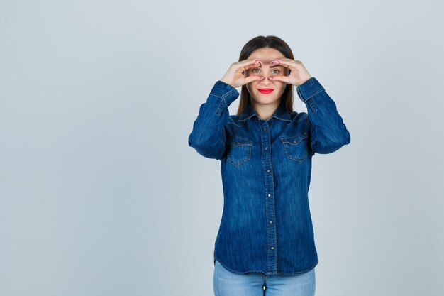 Young female in denim shirt and jeans pretending to peek through binoculars and looking cheery