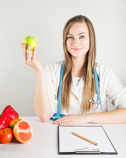 Young female dietician holding green apple with food and clipboard on desk