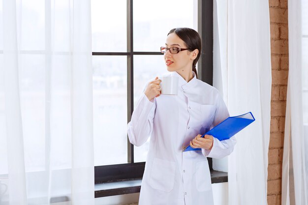 Young female doctor holding cup of coffee and binder near window. 