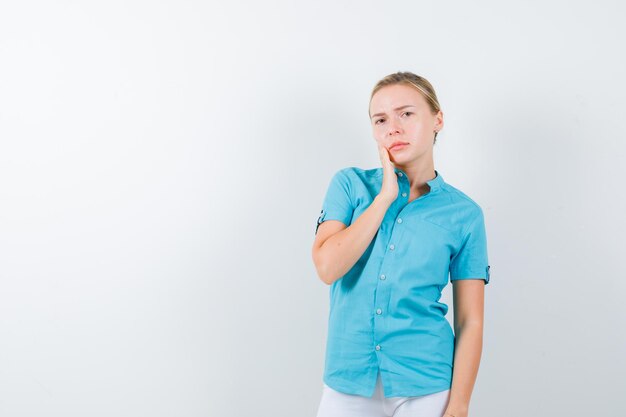 Young female doctor holding hand on cheek in medical uniform, mask and looking thoughtful