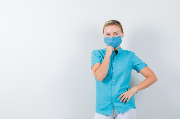 Young female doctor holding hand on chin while keeping hand on hip in medical uniform