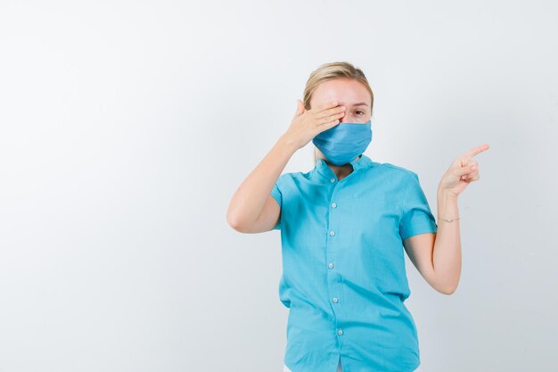 Young female doctor holding hand on eye while pointing to the right side isolated