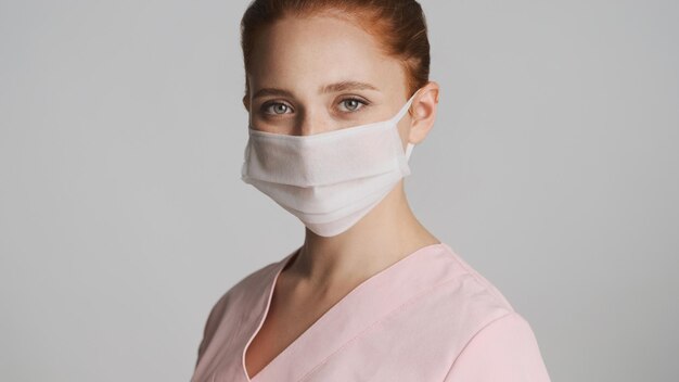 Young female doctor in medical mask happily looking in camera over white background Safety concept