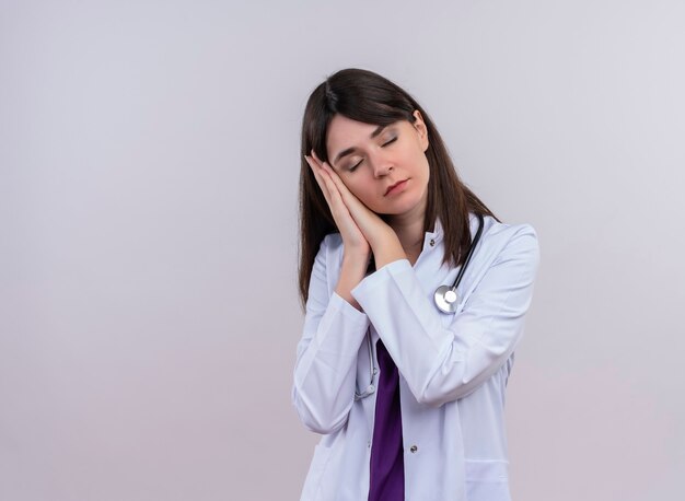 Young female doctor in medical robe with stethoscope pretends to sleep with closed eyes on isolated white wall