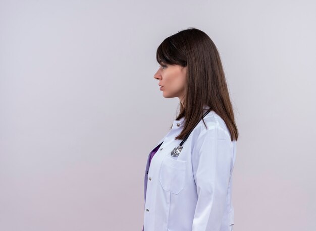 Young female doctor in medical robe with stethoscope stands sideways on isolated white wall