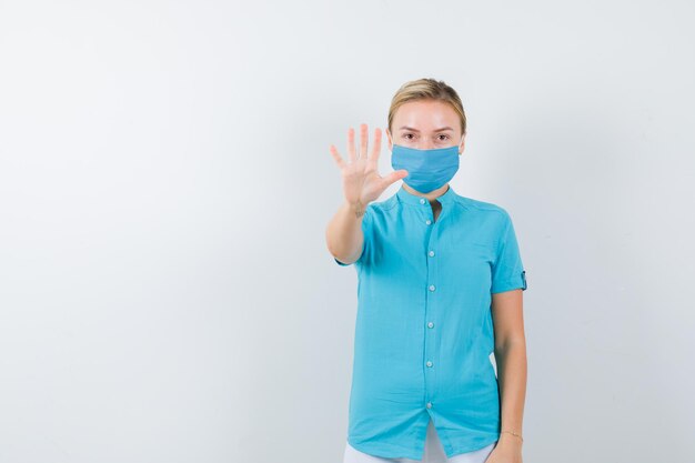 Молодая женщина-врач в медицинской форме, маска показывает жест стоп и выглядит серьезно