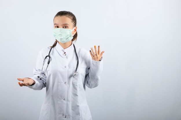 Giovane dottoressa in uniforme medica e maschera in piedi su sfondo bianco