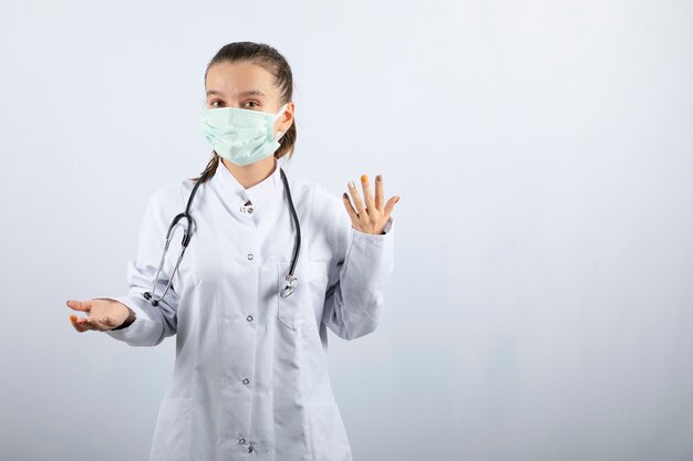 Молодая женщина-врач в медицинской форме и маске, стоя на белом фоне
