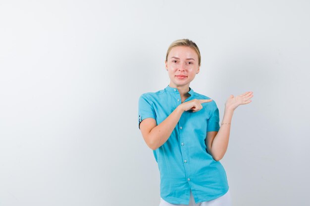 Young female doctor pointing at palm in medical uniform, mask and looking cheery